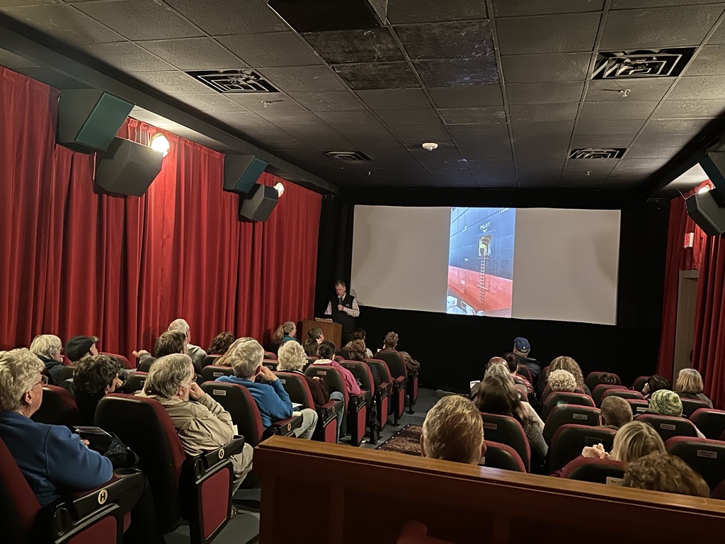 A man stands at a podium talking to an audience in a theater with a screen behind him.