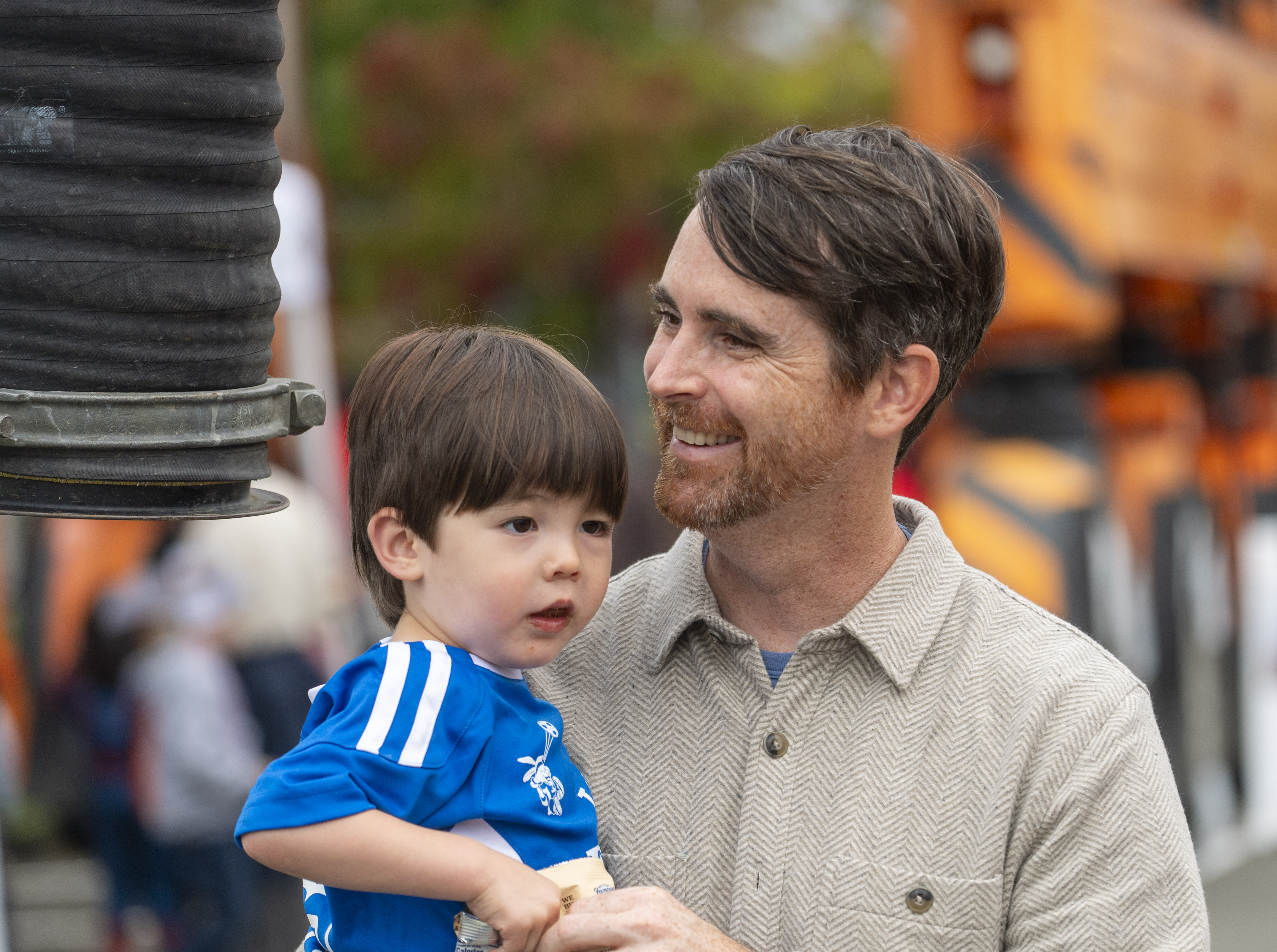 A man with brown hair smiles and holds a small child in a blue shrt.