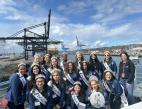 A group of young ladies in blue shirts and sashes pose in front of a cargo terminal.